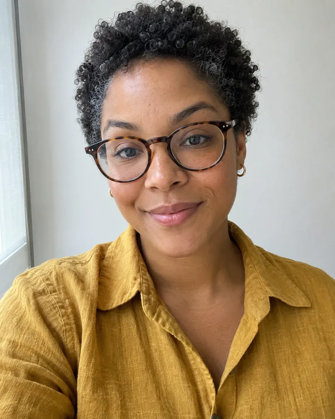 Photoreal synthetic selfie of a woman with short coiled hair, tortoiseshell glasses and mustard linen shirt