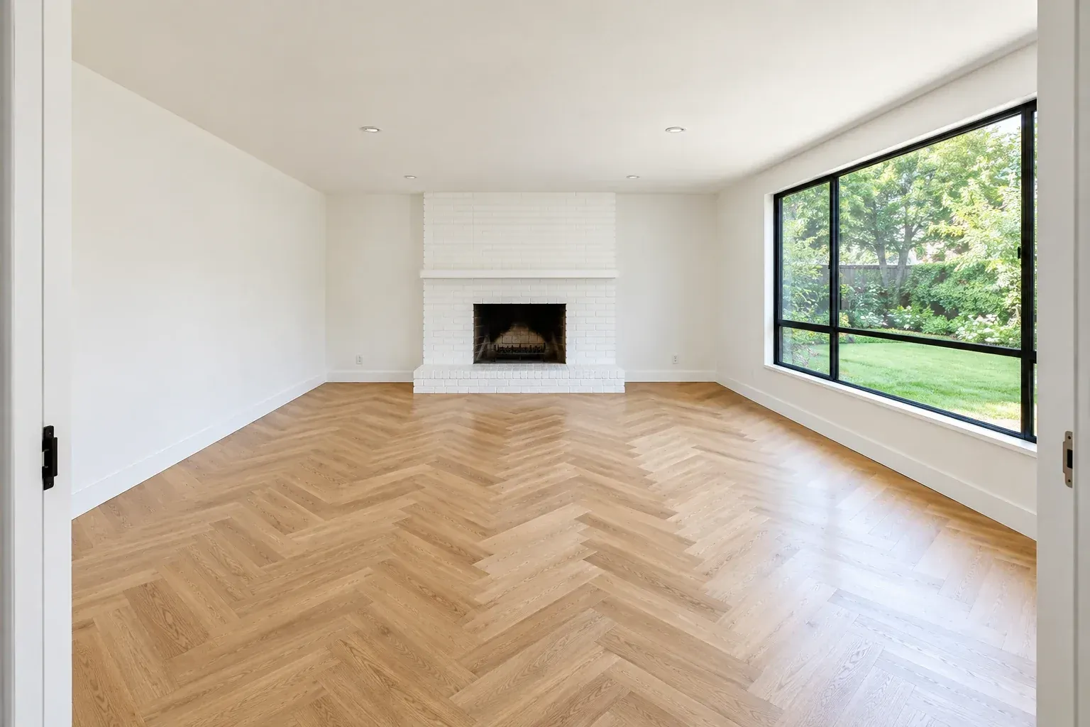 Empty mid-century living room with white walls, oak herringbone floor, black-framed window and white brick fireplace